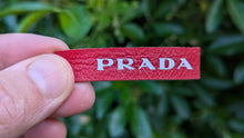 Load image into Gallery viewer, Red Prada tag held by a hand with a blurred green background