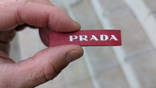 Load image into Gallery viewer, Red Prada tag held between fingers with a blurred background